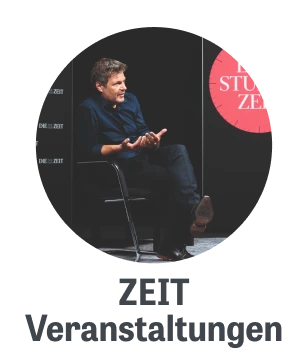 Mann in schwarzem Hemd sitzt auf einem Stuhl und gestikuliert während einer Diskussion auf einer Bühne, umgeben von Grafiken und dem Schriftzug "STUZEIT".