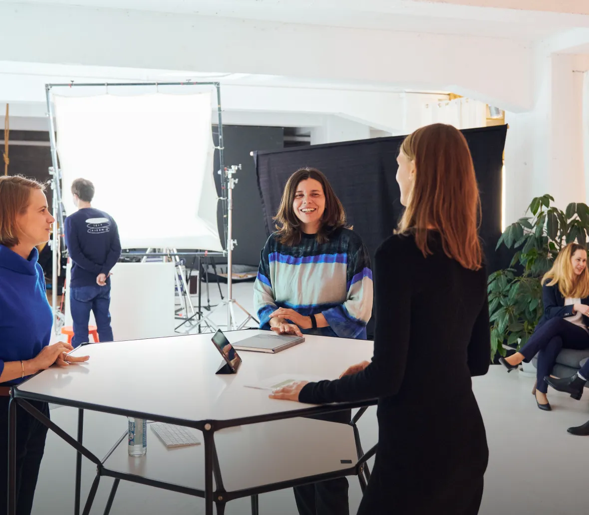 Drei Frauen unterhalten sich an einem Tisch in einem modernen, hellen Raum. Im Hintergrund ist eine Fotografie-Set mit einem großen Lichtpanel sichtbar. Eine Frau lächelt und trägt einen bunten Pullover, während die anderen in blauen und schwarzen Oberteilen gekleidet sind. Im Hintergrund sitzen weitere Personen, die an einem kreativen Arbeitsumfeld teilnehmen.