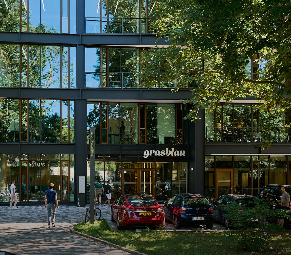 Moderne Bürogebäude mit großen Fenstern und dem Schriftzug "grasblau". Im Vordergrund sind mehrere Personen zu sehen, die das Gebäude betreten oder verlassen, sowie parkende Autos auf einem gepflasterten Parkplatz. Umgeben von Bäumen und Grünflächen.