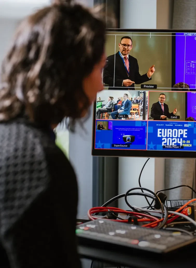 Eine Person sitzt vor einem Monitor, der mehrere Kameraperspektiven und eine Vorschau eines Sprechers auf einer Konferenz zeigt. Der Bildschirm zeigt Informationen zur Veranstaltung "EUROPE 2024", die am 19. und 20. März stattfindet. Im Vordergrund sind Kabel und ein Steuergerät sichtbar.