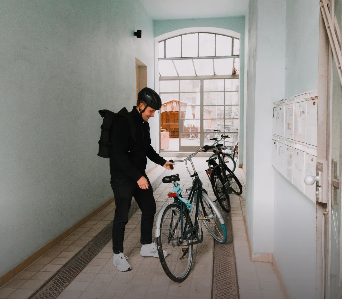 Junger Mann in einem Flur mit hellblauen Wänden, der ein Fahrrad mit einem Rucksack auf dem Rücken in einer Fahrradabstellanlage überprüft. Im Hintergrund sind weitere Fahrräder und Briefkästen sichtbar.